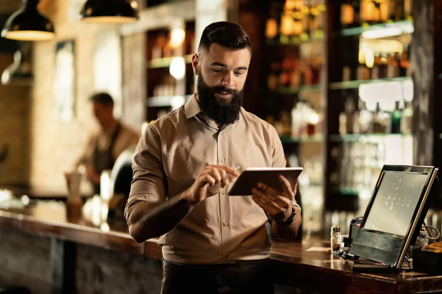 Hombre con barba y camisa social marrón en una cafetería, usando una tablet sobre la barra mientras hace un pedido o una consulta, con iluminación cálida y un mostrador al fondo.