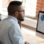 Hombre en una oficina, usando lentes, analiza un panel de gráficas e indicadores en una computadora con una interfaz de datos superpuesta en la pantalla, lo que sugiere análisis de rendimiento y estadísticas.