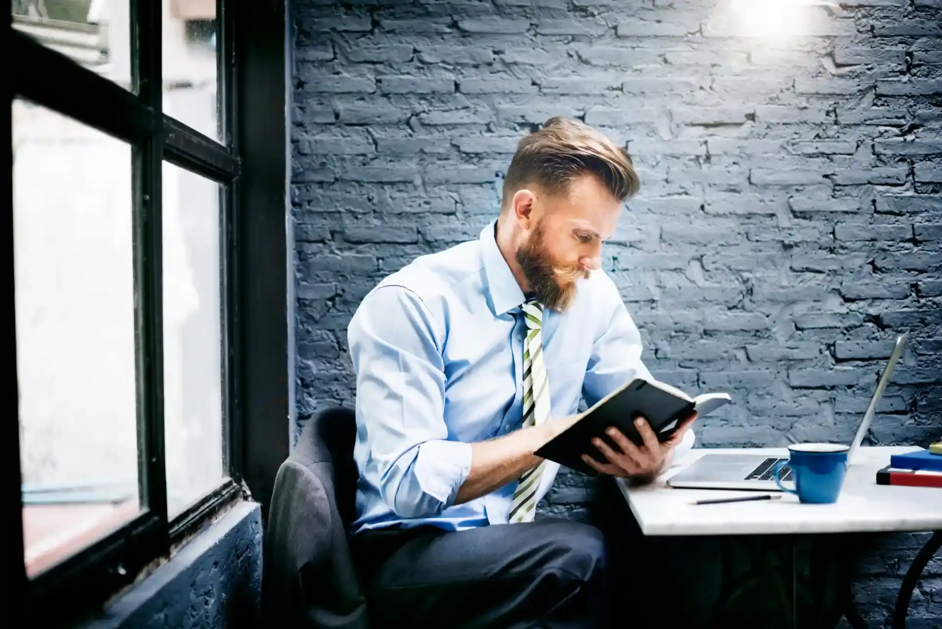 Hombre con camisa social azul y corbata a rayas sentado en una oficina con pared de ladrillo, usando una tablet y concentrado en la lectura, junto a una laptop y materiales de trabajo.