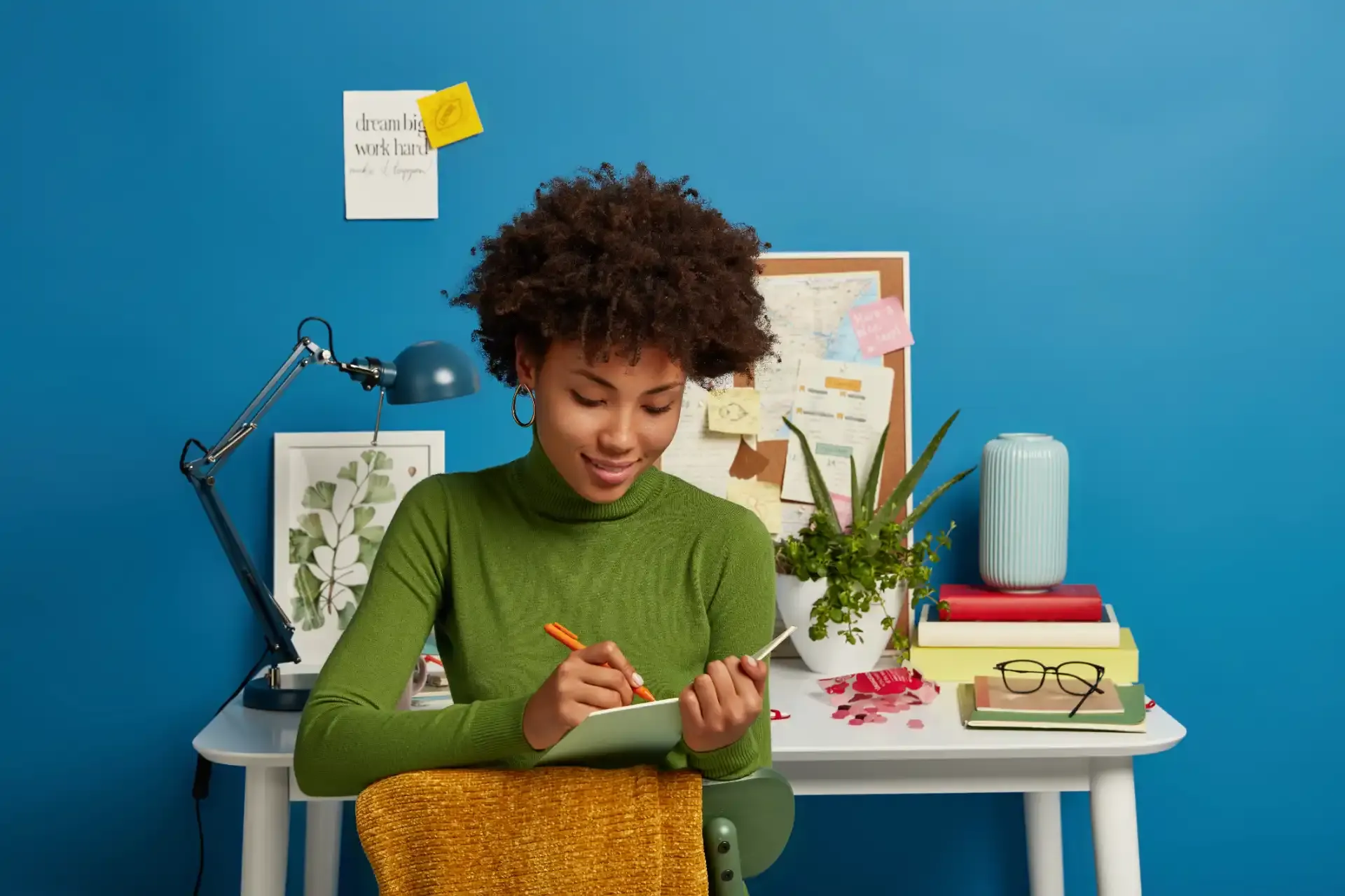 Joven sentada en una silla con asiento de tela natural, usando un suéter verde, dibujando en un bloc con un bolígrafo naranja en una mesa organizada con cuadernos, plantas y una lámpara azul frente a una pared azul.