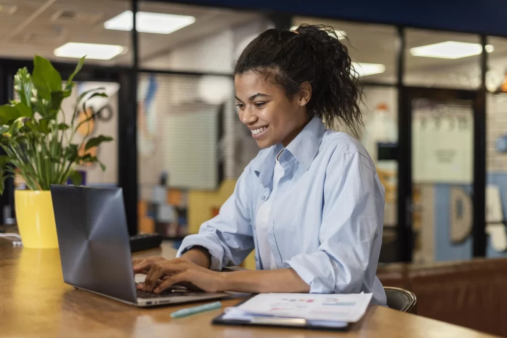 Mulher jovem e sorridente usando laptop em ambiente de escritório com plantas e iluminação adequada.