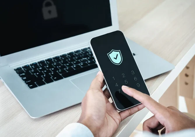 Pessoa segurando smartphone com ícone de proteção na tela, próximo a laptop, simbolizando segurança digital e proteção de dados online.