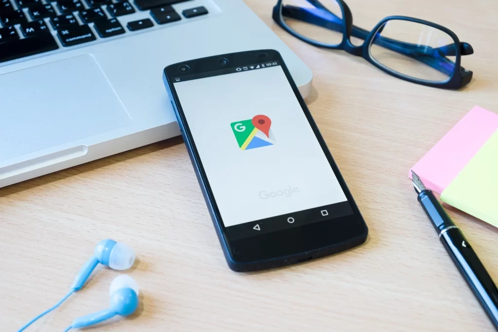 Smartphone com o aplicativo Google Maps aberto, ao lado de óculos, caderno, caneta e fones de ouvido, sobre uma mesa de trabalho.
