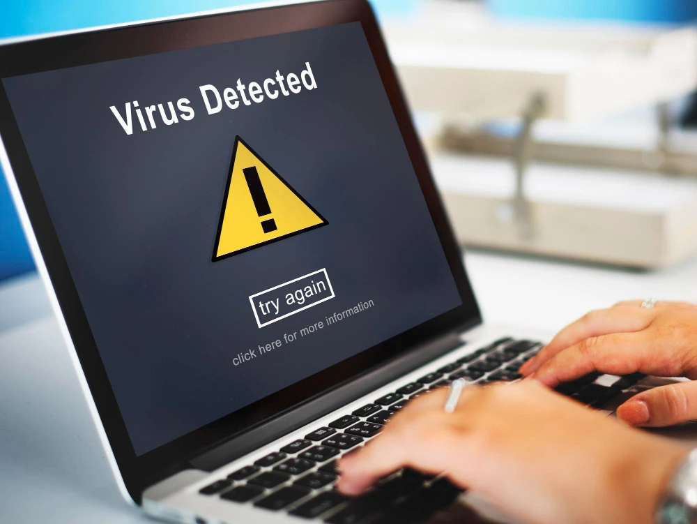 Pantalla de una laptop que muestra una alerta de “Virus Detected”, con un ícono de triángulo amarillo y un botón “try again”, mientras una persona escribe en el teclado, lo que sugiere una posible infección por malware.